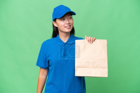 Foto de Young Asian woman taking a bag of takeaway food over isolated background looking to the side and smiling - Imagen libre de derechos