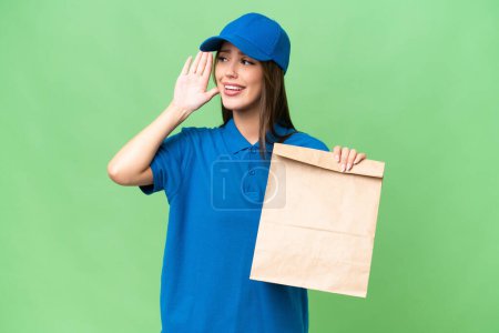 Téléchargez les photos : Young beautiful caucasian woman taking a bag of takeaway food over isolated background listening to something by putting hand on the ear - en image libre de droit