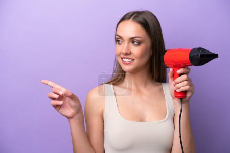 Téléchargez les photos : Young caucasian woman holding a hairdryer isolated on purple background pointing to the side to present a product - en image libre de droit