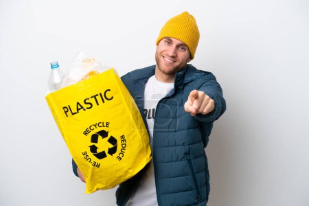 Foto de Young caucasian holding a bag full of plastic bottles to recycle isolated on white background pointing front with happy expression - Imagen libre de derechos