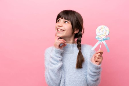 Foto de Little caucasian girl holding a lollipop thinking an idea while looking up - Imagen libre de derechos