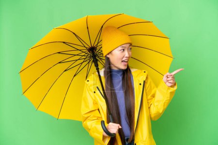 Téléchargez les photos : Young Asian woman with rainproof coat and umbrella over isolated chroma key background pointing finger to the side and presenting a product - en image libre de droit