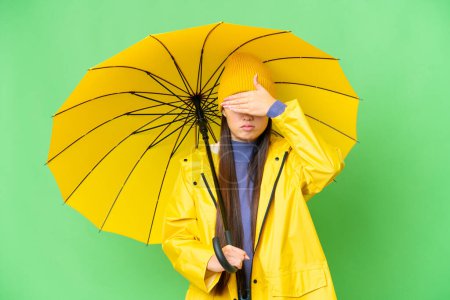 Téléchargez les photos : Young Asian woman with rainproof coat and umbrella over isolated chroma key background covering eyes by hands. Do not want to see something - en image libre de droit