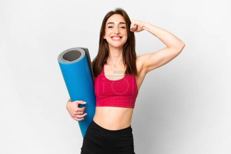 Young sport caucasian woman going to yoga classes while holding a mat over isolated white background doing strong gesture