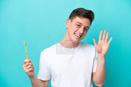 Foto de Joven brasileño cepillándose los dientes aislados sobre fondo azul saludando con la mano con expresión feliz - Imagen libre de derechos