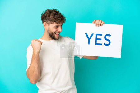 Young handsome caucasian man isolated on blue background holding a placard with text YES and celebration a victory