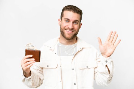 Foto de Young handsome blonde man holding a wallet over isolated white background saluting with hand with happy expression - Imagen libre de derechos