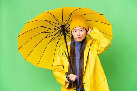 Téléchargez les photos : Young Asian woman with rainproof coat and umbrella over isolated chroma key background having doubts - en image libre de droit