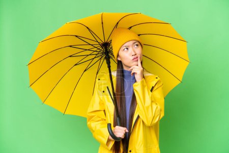 Téléchargez les photos : Young Asian woman with rainproof coat and umbrella over isolated chroma key background having doubts while looking up - en image libre de droit