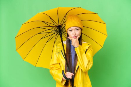 Téléchargez les photos : Young Asian woman with rainproof coat and umbrella over isolated chroma key background thinking - en image libre de droit
