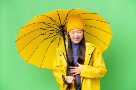 Téléchargez les photos : Young Asian woman with rainproof coat and umbrella over isolated chroma key background smiling a lot - en image libre de droit