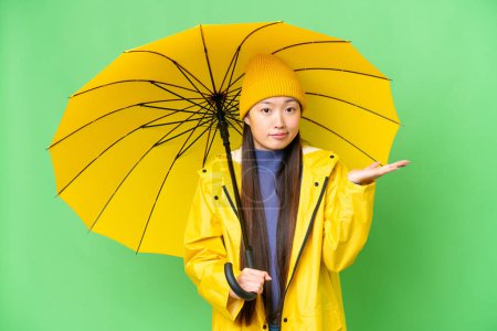 Téléchargez les photos : Young Asian woman with rainproof coat and umbrella over isolated chroma key background having doubts while raising hands - en image libre de droit