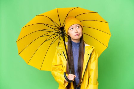 Téléchargez les photos : Young Asian woman with rainproof coat and umbrella over isolated chroma key background and looking up - en image libre de droit