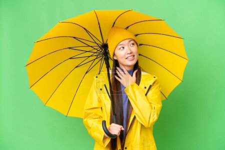 Téléchargez les photos : Young Asian woman with rainproof coat and umbrella over isolated chroma key background looking up while smiling - en image libre de droit