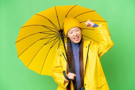 Téléchargez les photos : Young Asian woman with rainproof coat and umbrella over isolated chroma key background celebrating a victory - en image libre de droit
