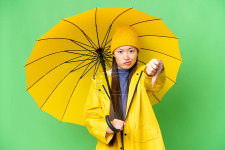 Téléchargez les photos : Young Asian woman with rainproof coat and umbrella over isolated chroma key background showing thumb down with negative expression - en image libre de droit