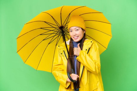 Téléchargez les photos : Young Asian woman with rainproof coat and umbrella over isolated chroma key background giving a thumbs up gesture - en image libre de droit