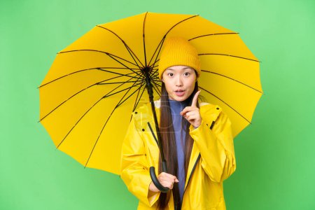 Téléchargez les photos : Young Asian woman with rainproof coat and umbrella over isolated chroma key background intending to realizes the solution while lifting a finger up - en image libre de droit