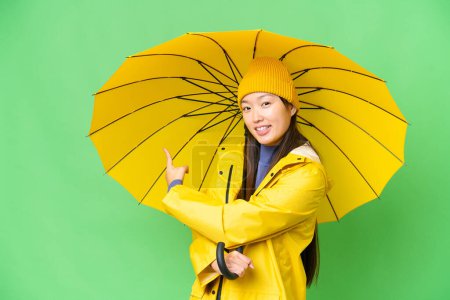 Téléchargez les photos : Young Asian woman with rainproof coat and umbrella over isolated chroma key background pointing back - en image libre de droit