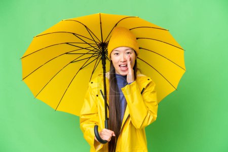 Téléchargez les photos : Young Asian woman with rainproof coat and umbrella over isolated chroma key background shouting with mouth wide open - en image libre de droit