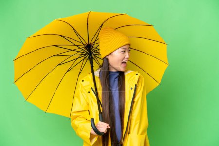 Téléchargez les photos : Young Asian woman with rainproof coat and umbrella over isolated chroma key background laughing in lateral position - en image libre de droit