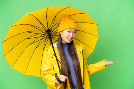 Téléchargez les photos : Young Asian woman with rainproof coat and umbrella over isolated chroma key background extending hands to the side for inviting to come - en image libre de droit