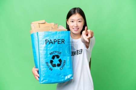 Foto de Young Asian woman holding a recycling bag full of paper to recycle over isolated chroma key background points finger at you with a confident expression - Imagen libre de derechos