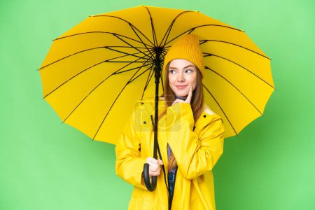 Téléchargez les photos : Young pretty woman with rainproof coat and umbrella over isolated chroma key background thinking an idea while looking up - en image libre de droit