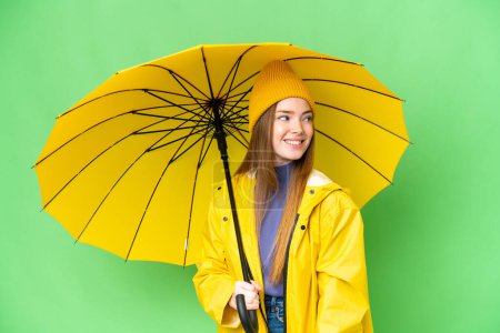 Téléchargez les photos : Young pretty woman with rainproof coat and umbrella over isolated chroma key background looking to the side and smiling - en image libre de droit