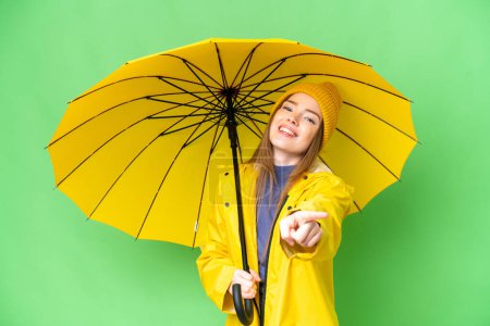Téléchargez les photos : Young pretty woman with rainproof coat and umbrella over isolated chroma key background pointing front with happy expression - en image libre de droit