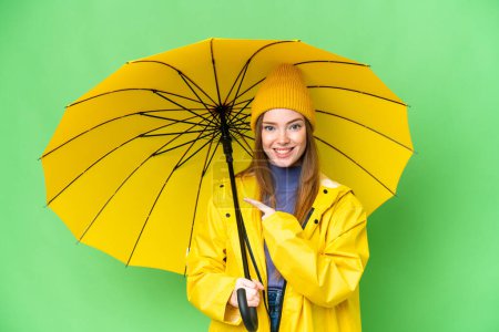 Téléchargez les photos : Young pretty woman with rainproof coat and umbrella over isolated chroma key background pointing to the side to present a product - en image libre de droit