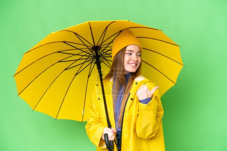 Téléchargez les photos : Young pretty woman with rainproof coat and umbrella over isolated chroma key background pointing to the side to present a product - en image libre de droit