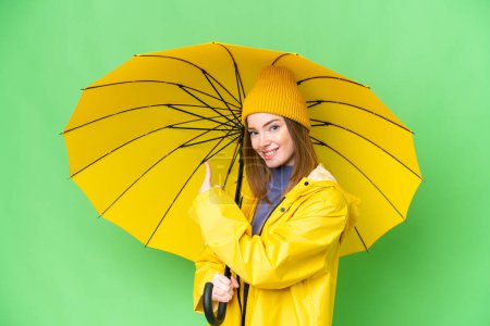 Téléchargez les photos : Young pretty woman with rainproof coat and umbrella over isolated chroma key background pointing back - en image libre de droit