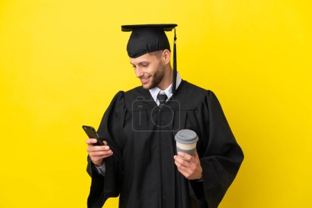 Photo pour Jeune diplômé universitaire caucasien isolé sur fond jaune tenant café à emporter et un mobile - image libre de droit