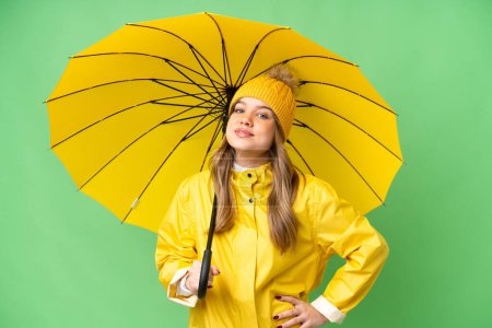 Téléchargez les photos : Jeune fille avec manteau imperméable et parapluie sur fond isolé chroma clé posant avec les bras à la hanche et souriant - en image libre de droit
