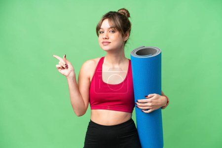 Foto de Joven chica deportiva que va a clases de yoga mientras sostiene una estera sobre un fondo de croma clave aislado apuntando con el dedo hacia un lado - Imagen libre de derechos