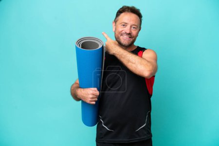 Foto de Hombre deportivo de mediana edad yendo a clases de yoga mientras sostiene una esterilla aislada sobre fondo azul apuntando hacia atrás - Imagen libre de derechos