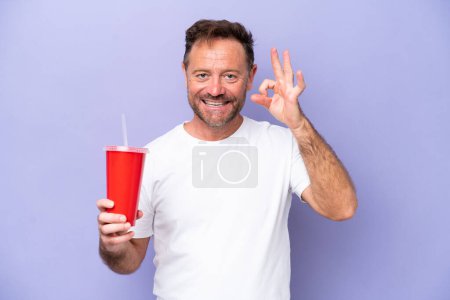 Foto de Hombre caucásico de mediana edad sosteniendo soda aislada sobre fondo púrpura mostrando signo aceptable con los dedos - Imagen libre de derechos