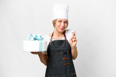 Photo for Young pastry blonde woman with a big cake over isolated white background with fingers crossing and wishing the best - Royalty Free Image