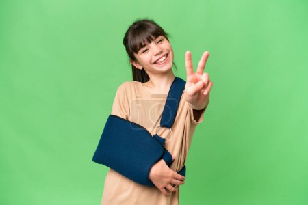 Photo for Little caucasian girl with broken arm and wearing a sling over isolated background smiling and showing victory sign - Royalty Free Image