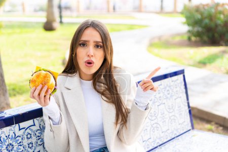 Young pretty woman holding a burger at outdoors surprised and pointing side