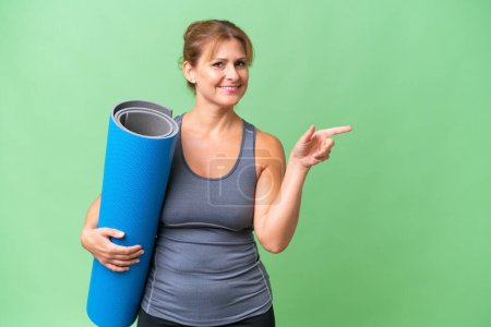 Foto de Mujer deportiva de mediana edad que va a clases de yoga mientras sostiene una esterilla sobre un fondo aislado apuntando con el dedo hacia un lado - Imagen libre de derechos