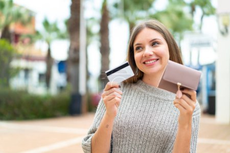 Foto de Joven mujer rubia bonita sosteniendo billetera y tarjeta de crédito con expresión feliz - Imagen libre de derechos