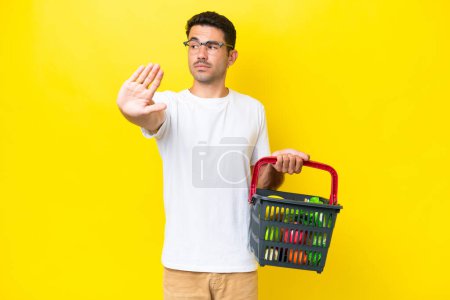 Foto de Joven hombre guapo sosteniendo una cesta llena de comida sobre fondo amarillo aislado haciendo gesto de parada y decepcionado - Imagen libre de derechos