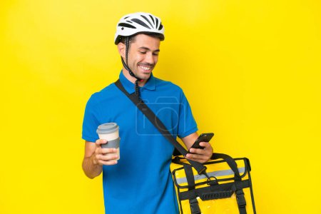 Téléchargez les photos : Jeune homme beau avec sac à dos thermique sur fond jaune isolé tenant café à emporter et un mobile - en image libre de droit