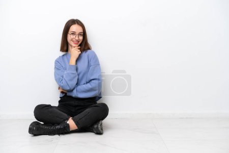 Téléchargez les photos : Jeune femme ukrainienne assise sur le sol isolé sur fond blanc avec des lunettes et souriant - en image libre de droit