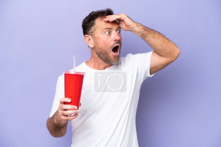 Foto de Hombre caucásico de mediana edad sosteniendo soda aislada sobre fondo púrpura haciendo gesto sorpresa mientras mira a un lado - Imagen libre de derechos