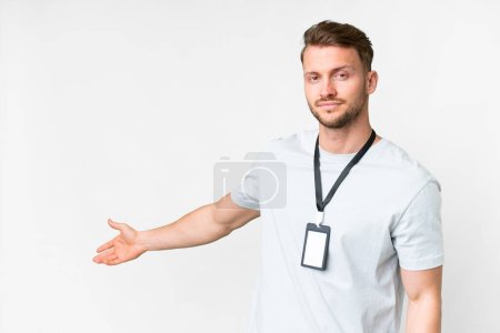 Foto de Joven hombre caucásico con tarjeta de identificación sobre fondo blanco aislado extendiendo las manos a un lado para invitar a venir - Imagen libre de derechos