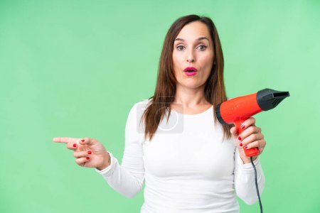 Photo for Middle age woman holding a hairdryer over isolated chroma key background surprised and pointing side - Royalty Free Image
