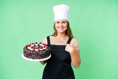 Foto de Mujer pastelera de mediana edad sosteniendo una gran torta sobre un fondo aislado con expresión facial sorpresa - Imagen libre de derechos